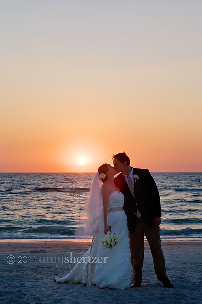 The bride and groom kiss on the beach as the sun sets over the water.